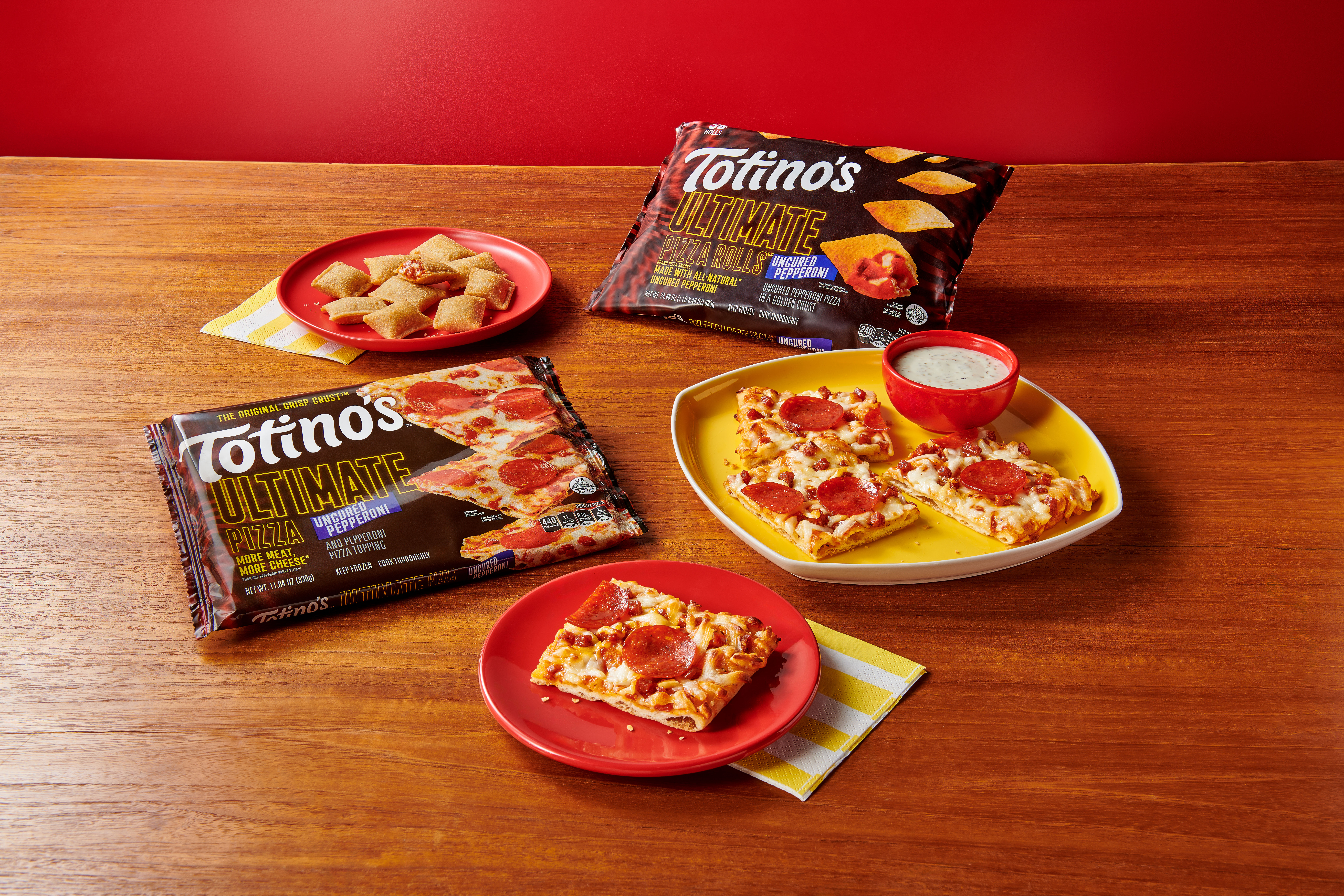 Package of Totino's Ultimate Pizza and Totino's Ultimate Pizza Rolls on a wooden table alongside red and yellow plates with cooked pizza rolls and square slices of pizza with a side of ranch in a red cup.